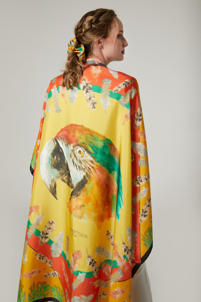 Mujer elegante con coletero en trenza y pañuelo de seda natural pura grande como chal, color naranja y amarillo con diseños de plumas y loro
