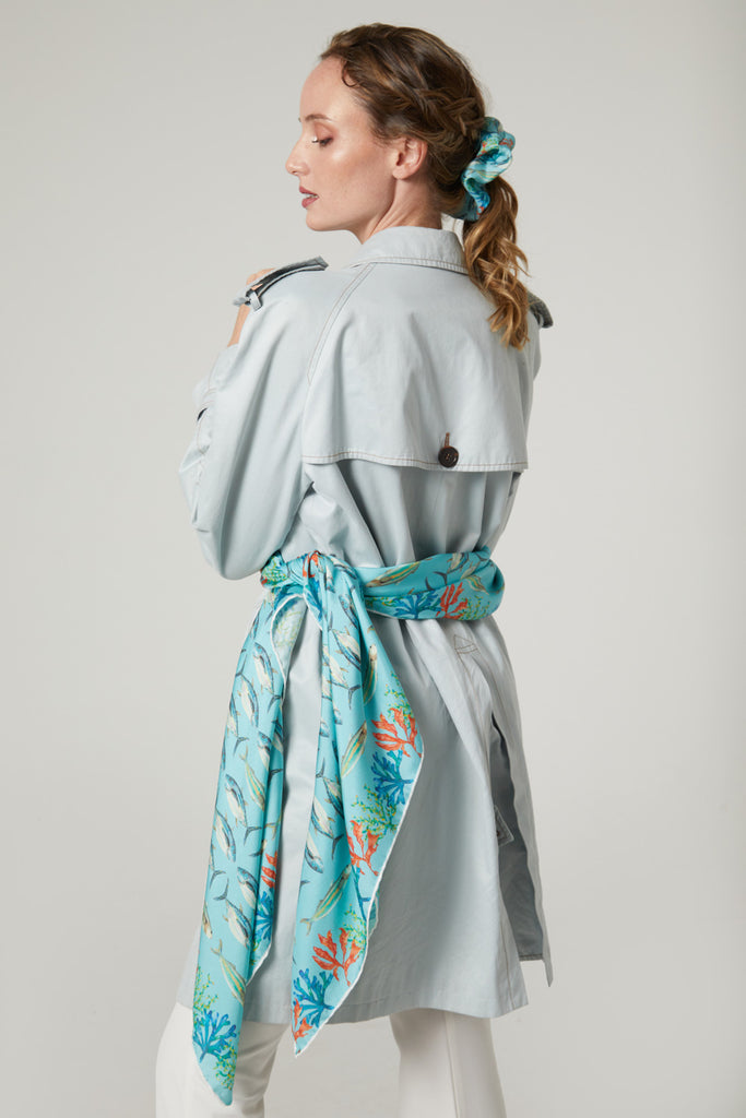 Mujer elegante de pie con gabardina y pañuelo de seda natural pura maxi azul claro atado a la cintura con coletero de la misma colección Martsen - Medas